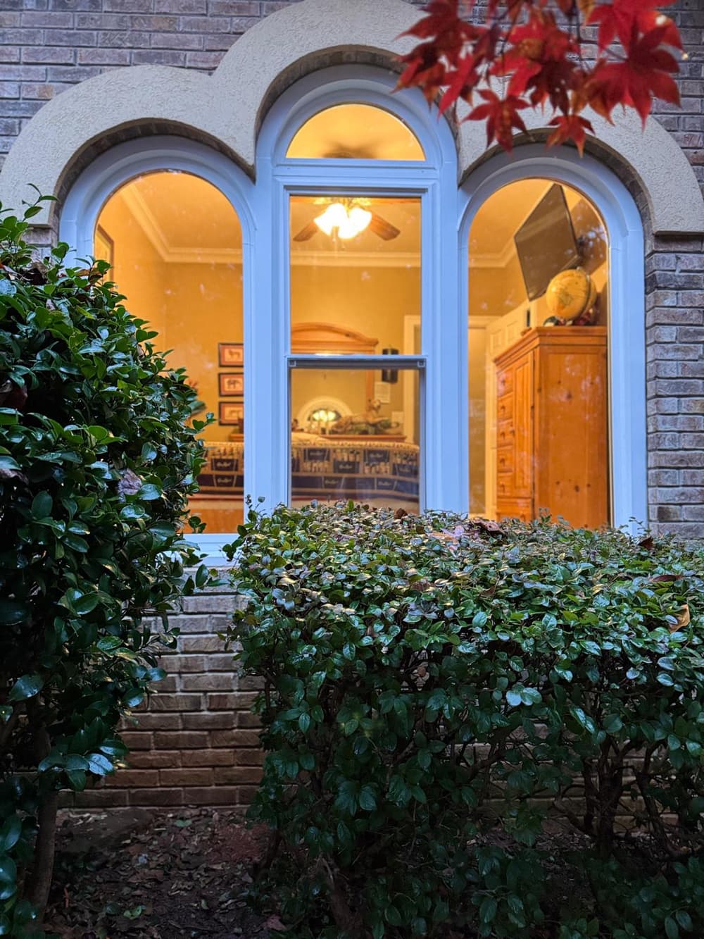 Three arched windows glowing warm at dusk behind a hedge